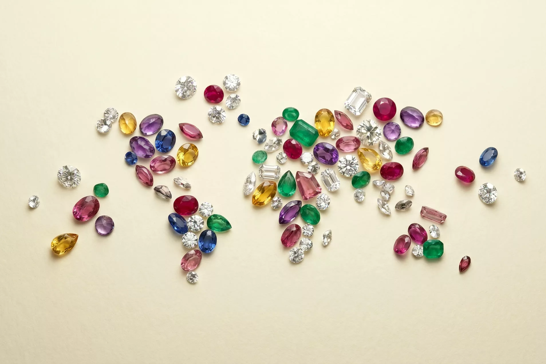 Polished diamonds and faceted multi-color gemstones (rubies, sapphires, emeralds, citrines, amethysts, pink tourmaline) scattered on warm cream paper under soft studio light